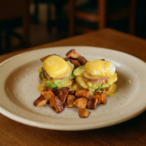 Two eggs Benedict with avocado, bacon, and hollandaise sauce, served with roasted potatoes.